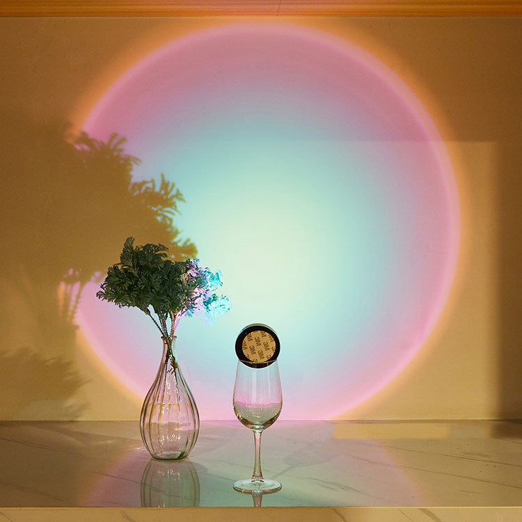 Vase with greenery and a glass of wine on a reflective surface with a colorful circular light projection on the wall.